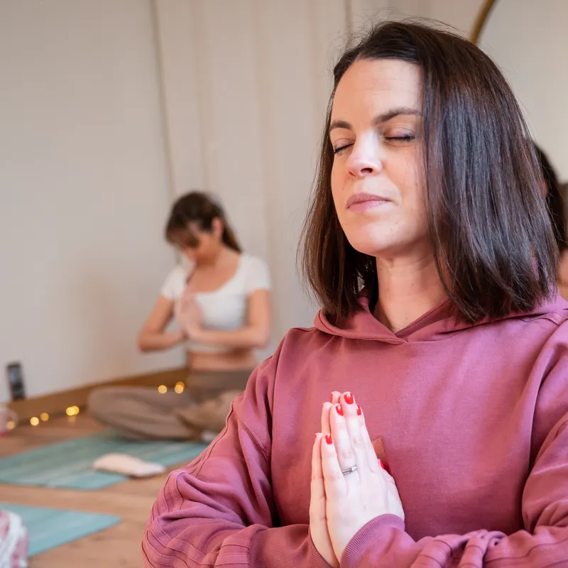 Élève assise en tailleur, les yeux fermés et les mains jointes devant la poitrine, en sweat à capuche rose ; une autre participante floue en arrière-plan.
