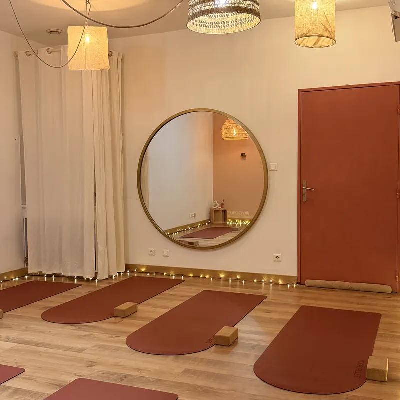 Salle du studio vue de face, avec un grand miroir rond à cadre doré, une porte terracotta, des rideaux blancs et des tapis alignés sur le parquet.