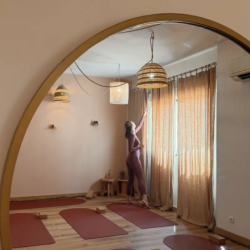 Reflet dans un miroir rond à cadre doré : une personne debout ferme un rideau, sous une suspension en fibres naturelles.