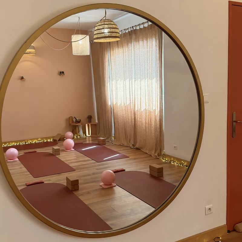 Grand miroir rond à cadre doré reflétant la salle : rideaux beiges, tapis terracotta et balles de Pilates roses au sol.