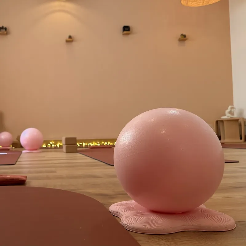 Vue à hauteur de sol de la salle du studio : au premier plan, une balle de Pilates rose sur son support, avec des tapis, des briques en liège et des guirlandes lumineuses au fond.
