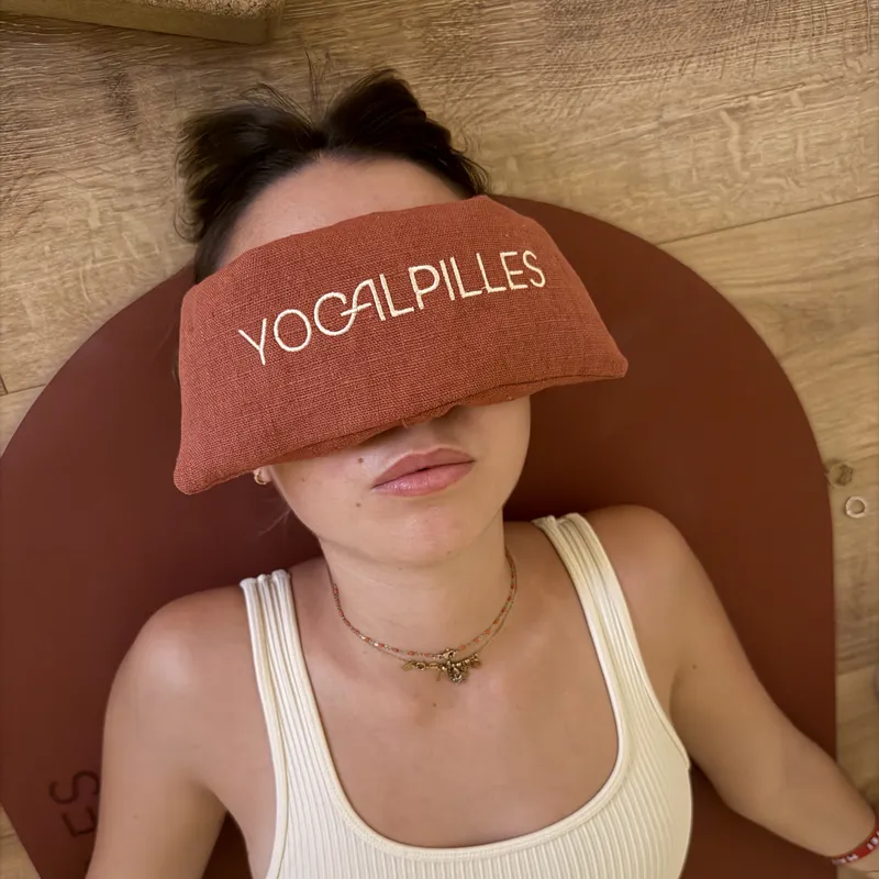 Personne allongée sur un tapis terracotta, un coussin pour les yeux brodé « Yogalpilles » posé sur le visage, en débardeur côtelé crème.