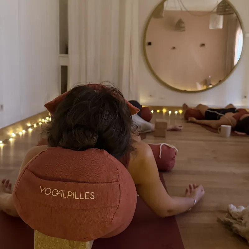 Séance de relaxation dans une salle claire : une personne vue de dos, allongée sur un tapis avec un bolster brodé « Yogalpilles » posé sur une brique en liège, d'autres élèves au fond, devant un grand miroir rond.