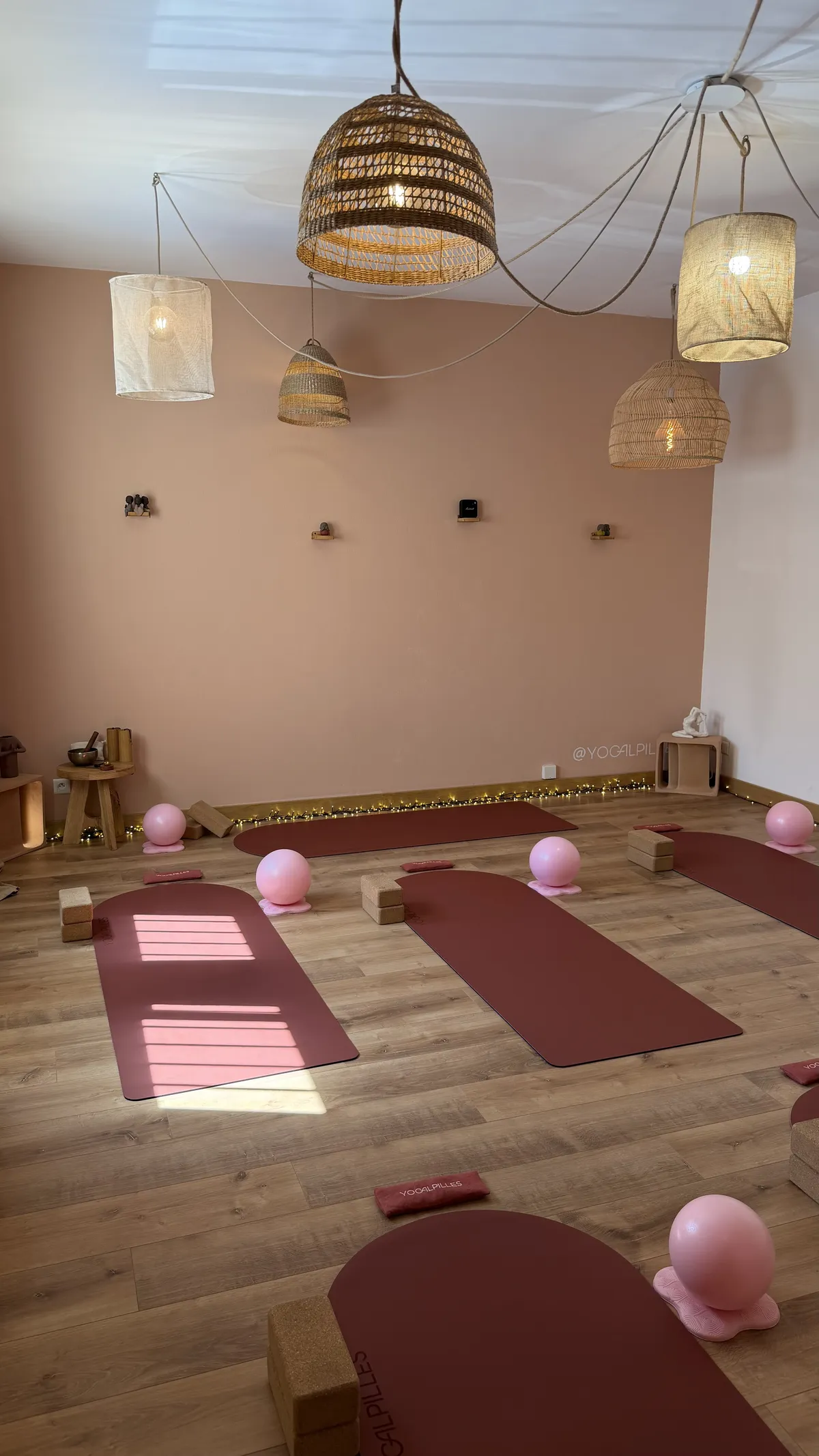 Salle de pratique du studio Yogalpilles, aux murs rose pâle, avec de grands rideaux beiges, des tapis terracotta et des briques en liège.00