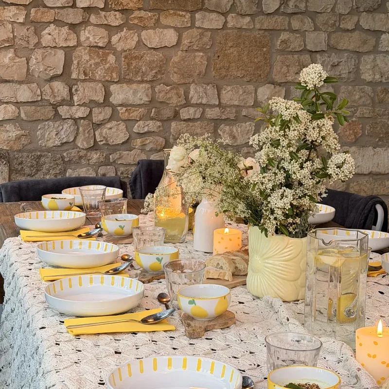 Table fleurie dressée pour un repas, avec bouquet de fleurs blanches, assiettes et verres, devant un mur en pierre