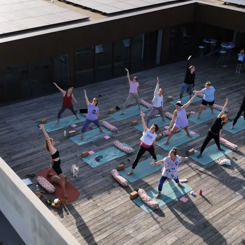 Vue de dessus d’un cours collectif sur une terrasse en bois, élèves debout bras levés sur des tapis turquoise