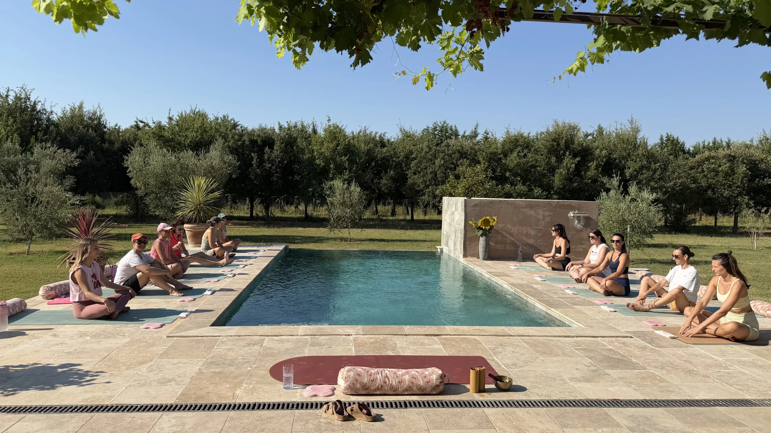 Cours de yoga en extérieur : élèves assises sur des tapis de chaque côté d’une piscine, entourée d’arbres, sous un ciel bleu