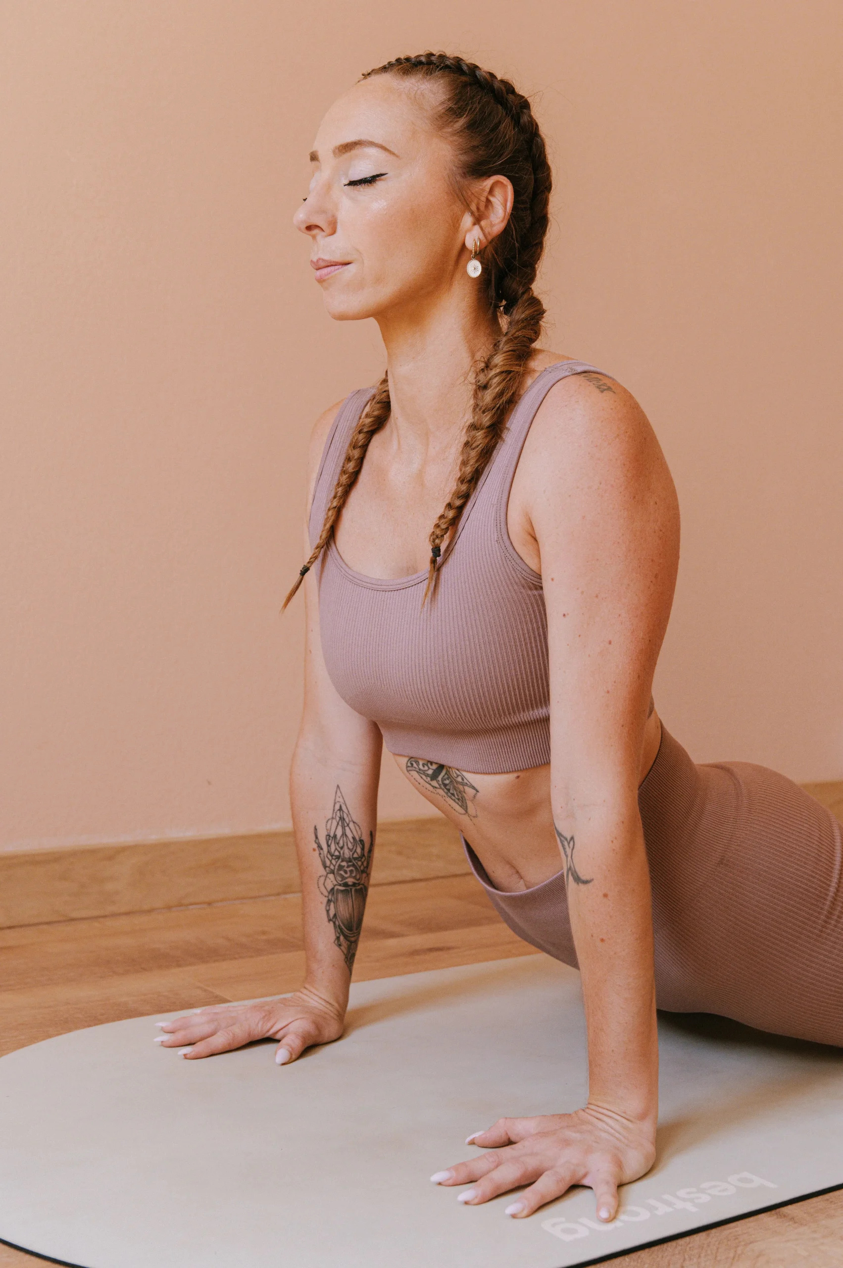Charlotte avec de longues tresses en posture du cobra, yeux fermés, sur un tapis de yoga gris clair devant un mur rose