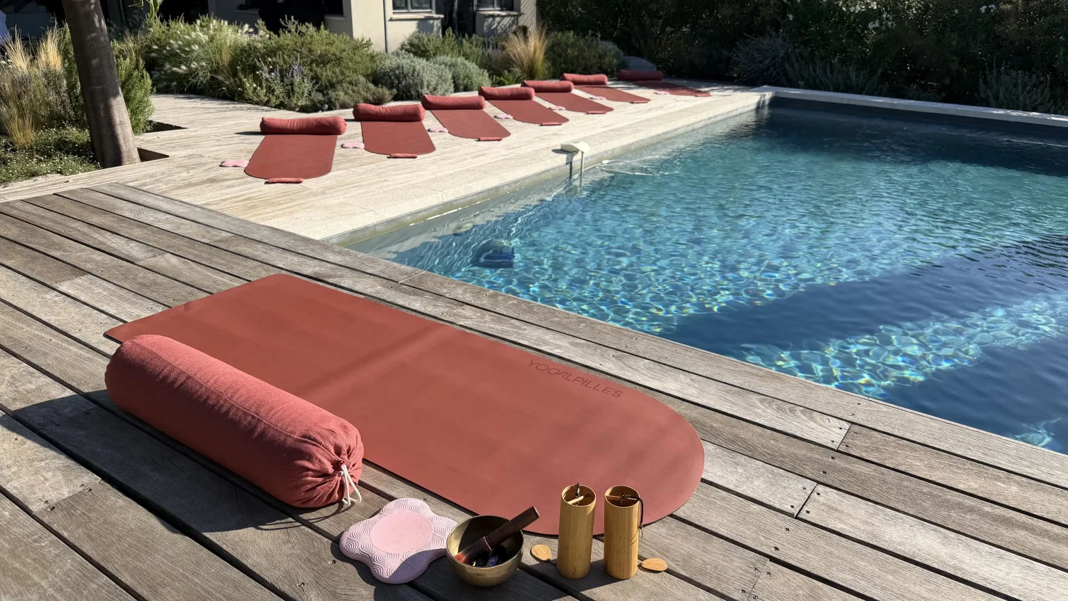 Tapis terracotta, bolster, bol tibétain et instruments en bambou au bord d'une piscine, d'autres tapis alignés au fond